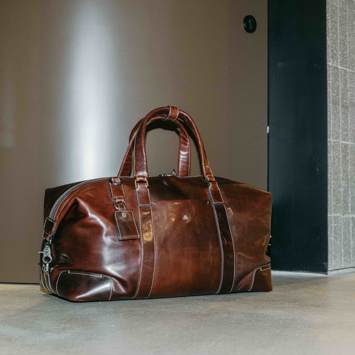 Mocha leather Weekender standing on a concrete floor beside a doorway