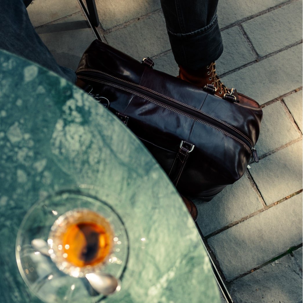 Mocha leather Weekender on a pavement beside a café table and a glass of whisky