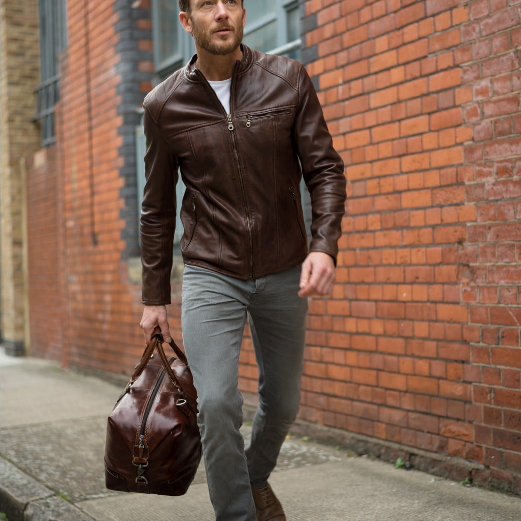 Man in a leather jacket walking past a brick wall carrying the Mocha leather Weekender
